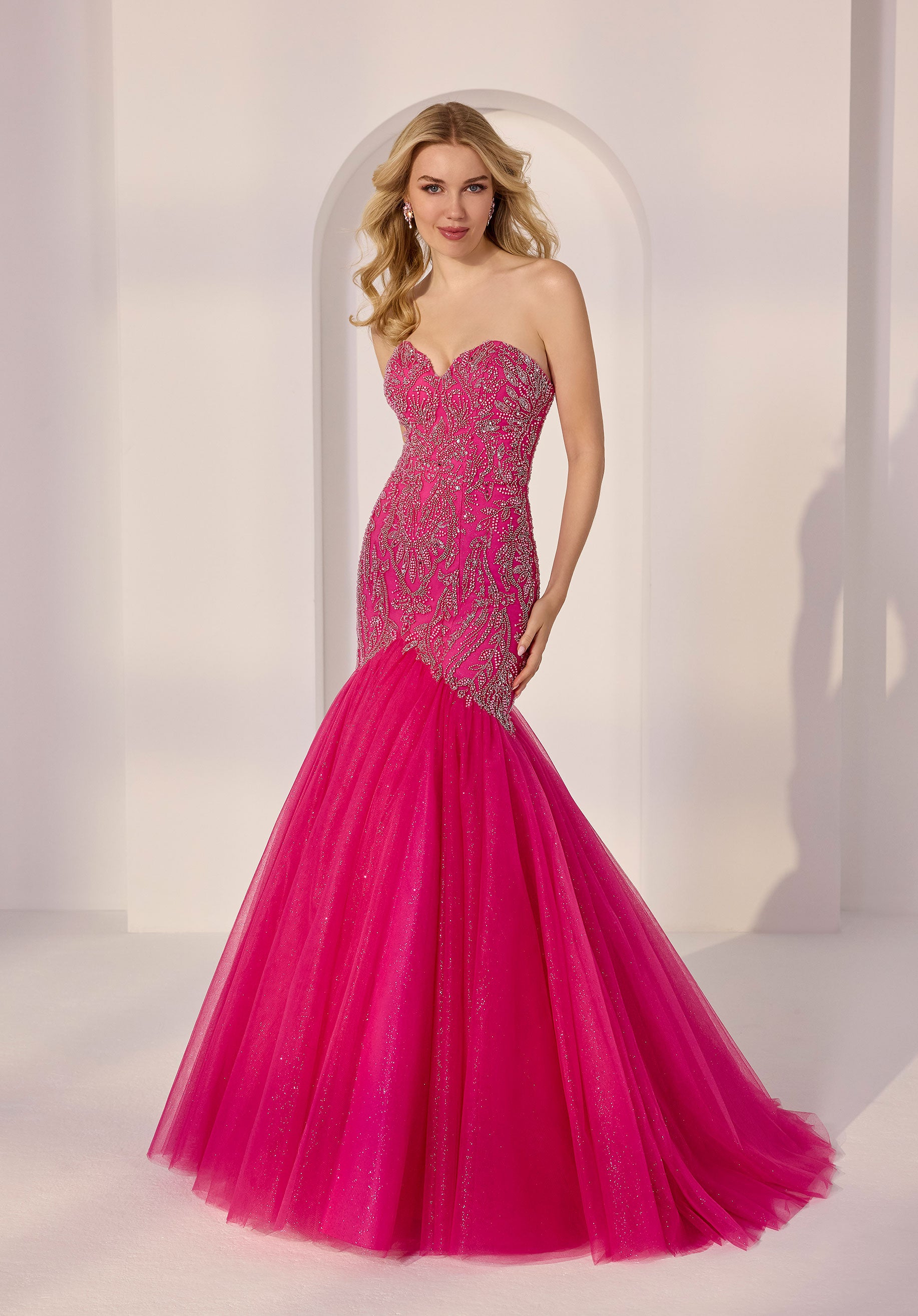 A woman stands against a light-colored studio backdrop, wearing the Morilee 49203 strapless mermaid prom dress with a bright pink tulle skirt and an intricate sequin bodice.